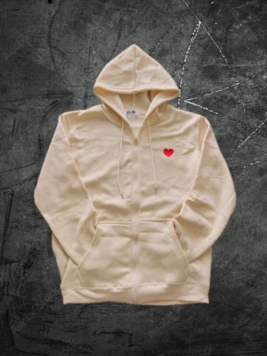 Comme des Garçons Zip-up hoodie - Snygg beige hoodie från Comme des Garçons PLAY med dragkedja, huva och fickor framtill. På bröstet sitter det ikoniska röda hjärtat med ögon. Tillverkad i mjuk bomull som känns skön mot huden. Perfekt för en chill och stilren look.