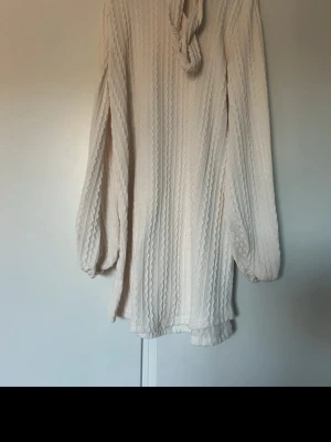 Krämvit stickad tröja med knytband - Säljer en krämvit stickad tröja med snygg kabelstickning och knytband vid halsen. Tröjan har långa, lite puffiga ärmar och avslappnad passform. Perfekt för dig som gillar en stilren och mysig look.