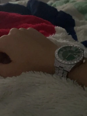 Silverfärgad klocka med grönt ur - Förminskad Stilren klocka med grönt ur och romerska siffror. Armbandet och boetten är silverfärgade och dekorerade med gnistrande stenar för en extra lyxig känsla. Perfekt accessoar för dig som vill sticka ut.