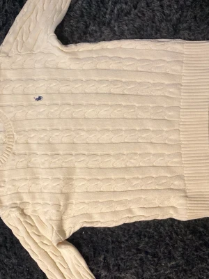 Vit stickad Ralph Lauren Tröja - Kabelstickad tröja i offwhite från Polo Ralph Lauren med klassisk blå logga broderad på bröstet. Tröjan har rund halsringning, ribbade muddar och nederkant. Perfekt för dig som gillar tidlös och stilren design.