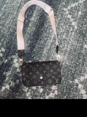 Louis Vuitton monogram axelväska - Säljer en snygg axelväska från Louis Vuitton med klassiskt monogrammönster i brunt och beige. Väskan har en bred, ljusrosa axelrem med logotyp och guldfärgade detaljer. Perfekt för dig som vill ha en trendig och lyxig accessoar.