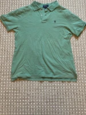 Grön pikétröja från Polo Ralph Lauren - Klassisk grön pikétröja från Polo Ralph Lauren med korta ärmar och krage. Tröjan har två knappar vid halsen och den ikoniska mörkblå logotypen broderad på bröstet. Tillverkad i mjuk bomull som känns skön mot huden. Den är i storlek L i 14-16 år men skulle säga att den sitter som en S i herrkläder 
