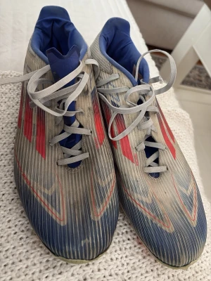 Adidas fotbollsskor blå/röd - Säljer ett par Adidas fotbollsskor med blå och röda detaljer samt vita snören. Skorna har en strömlinjeformad design och ovandel i syntetmaterial. Insidan är blå och skon har klassiska Adidas-ränder på sidorna. Perfekta för dig som vill sticka ut på planen.