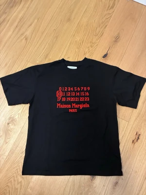 Maison Margiela svart t-shirt - Svart t-shirt från Maison Margiela med rött broderat tryck framtill som visar siffror och logotypen 'Maison Margiela PARIS'. Klassisk passform. Perfekt för dig som gillar streetwear och exklusiva märken.