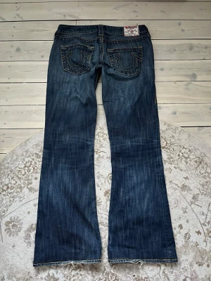 True Religion mörkblå bootcut jeans - Säljer ett par mörkblå lowrise bootcut jeans från True Religion med kontrastsömmar och ikoniska bakfickor med broderad logga.  Midja: 38cm Innerbenslängd: 76cm