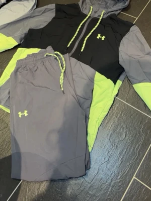 Under Armour träningsset grå/svart - Snyggt träningsset från Under Armour med hoodie och byxor.Jackan är i storlek S men passar M och byxorna är M. Setet är i grått, svart och neongult med matchande snören och logga på både tröja och byxa. Tillverkat i lätt och ventilerande material, perfekt för träning eller chill.