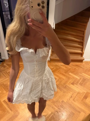 Vit kort klänning med spetsdetaljer - Supersöt vit kort klänning med volang och spetsdetaljer vid axelband och nederkant. Klänningen har knappar framtill och en figurnära passform med markerad midja. Perfekt för sommarens alla tillfällen!