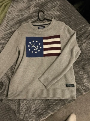 Grå stickad tröja från Sebago - Säljer en grå stickad tröja från Sebago med ett stort flaggliknande mönster på bröstet i blått, vitt och vinrött. Tröjan har rund halsringning, ribbade muddar och en patch med Sebago-logga vid nederkanten. Perfekt för dig som gillar klassisk stil.