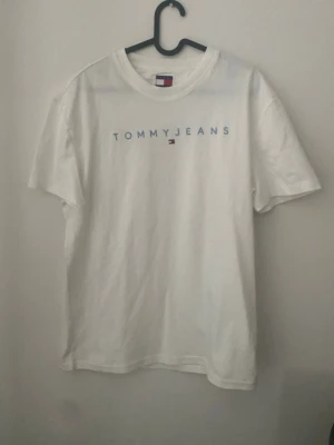 Vit T-shirt från Tommy Jeans - Vit T-shirt från Tommy Jeans med blå text och den klassiska loggan på bröstet. T-shirten har rund halsringning och korta ärmar. Tillverkad i mjuk bomull för en skön och avslappnad stil. Skriv för mått. Pris kan diskuteras