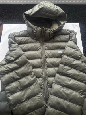 Olivgrön pufferjacka från Levi's - Snygg olivgrön pufferjacka från Levi's med huva och dragkedja framtill. Jackan har quiltad design med breda kanaler och en diskret logotyp på bröstet. Perfekt för kalla dagar och riktigt skön att bära tack vare det vadderade materialet.