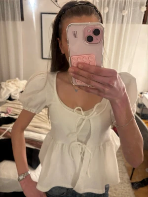 Vit blus med puffärm och knyt - Supersöt vit blus med puffärmar och två knyt framtill. Blusen har en lätt rynkad midja och en feminin, luftig passform. Perfekt att styla med jeans eller kjol. Aldrig använd och aldrig haft användning av.
