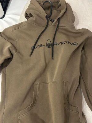 Beige hoodie från Sail Racing - Snygg beige hoodie från Sail Racing med svart logga tryckt över bröstet. Hoodien har huva med svarta dragsnören och en stor magficka framtill. Perfekt för en avslappnad och sportig stil.