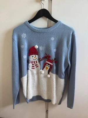 Blå jultröja med snögubbe och pingvin - Mysig ljusblå stickad tröja med snögubbe och pingvin i julmotiv framtill. Tröjan har vita snöflingor, röda detaljer och ribbade muddar vid ärmslut och nederkant. Perfekt för vinterns alla tillfällen!