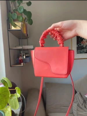 Korallrosa ATP Atelier handväska - Säljer en snygg korallrosa handväska från ATP Atelier med unik scrunchie-handtag och avtagbar axelrem. Väskan är tillverkad i slätt skinn och har guldfärgade metalldetaljer. Designen är stilren med en asymmetrisk lock och kompakt form, perfekt för att lyfta din look.