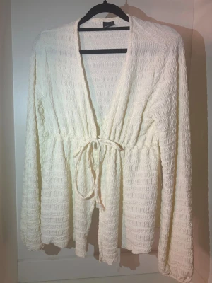 Vit strukturerad kofta med knytband - Säljer en vit kofta med vågig struktur och knytband i midjan. Koftan har lång ärm, djup V-ringning och ett mjukt, stretchigt material. Perfekt att slänga över en topp för en chill look.