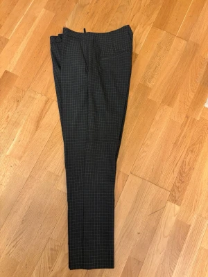 Mörkgrå rutiga kostymbyxor - Säljer ett par mörkgrå kostymbyxor med tunna vita rutor i ett klassiskt rutmönster. Byxorna har rak passform och pressveck framtill. Perfekta för en stilren och trendig look. Materialet känns mjukt och följsamt.