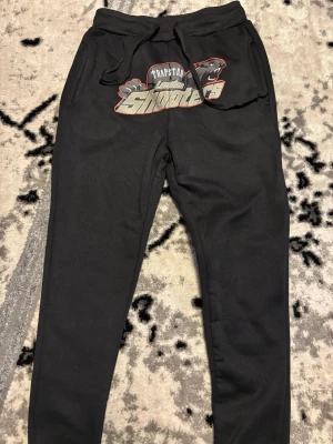 Trapstar London svarta mjukisbyxor - Svarta mjukisbyxor från Trapstar London med stort tryck på framsidan där det står 'Shooters'. Byxorna har dragsko i midjan och muddar vid bensluten. Perfekta för en avslappnad och street-inspirerad stil.