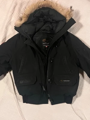 Svart Canada Goose pufferjacka med päls - Snygg svart pufferjacka från Canada Goose med fuskpäls. Jackan har stora fickor framtill, dragkedja och knappar samt ribbade muddar vid ärmslut och midja. Perfekt för kalla vinterdagar då den är riktigt varm. Har själv inte använt den lika mycket som jag trott, men den har lite syn på användning då jag är den andra ägaren av jackan. Pris går att diskutera, köpte den själv för 4000kr.