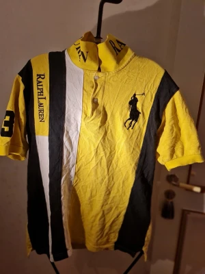 Gul och svart piké från Ralph Lauren - Snygg pikétröja i gult, svart och vitt från Ralph Lauren med stora logotryck, klassisk krage och knappar framtill. Tröjan har korta ärmar, nummer 3 på ärmen och den ikoniska polospelaren broderad på bröstet. Perfekt för en sportig och trendig look.
