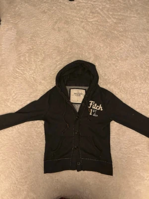 Mörkgrå hoodie från Abercrombie & Fitch - Mörkgrå hoodie från Abercrombie & Fitch med broderad text 'Fitch 1st div' på bröstet. Hoodien har knappar framtill, dragsko i huvan och två fickor. Tillverkad i mjukt bomullsmaterial och har en fast passform. Pris kan diskuteras hör av er vid minsta fundering😁😁🙌🙌