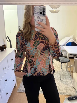 Blommig peplumblus med trekvartsärm - Snygg blus med peplum-modell och färgglatt blommönster i rosa, turkos, lila och beige toner. Blusen har rund halsringning och trekvartsärmar. Perfekt för dig som vill ha en färgklick i garderoben! Passar M/L