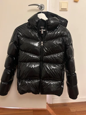 Svart glansig pufferjacka med huva - Snygg svart jacka från Colmar som är glansig. Jackan är använd endast ett få tal gånger förra vintern då den köptes ny. Skulle säga den är 34/36. Köptes ny för 4999kr
