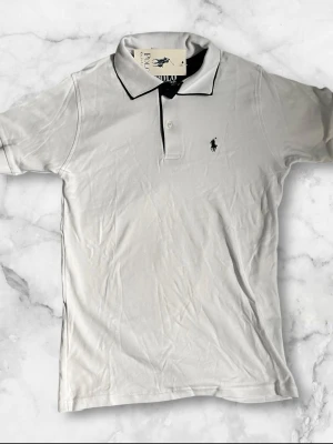 Vit pikétröja från Ralph Lauren - Klassisk vit pikétröja från Ralph Lauren med svart logga broderad på bröstet och svart detalj på kragen. Tröjan har korta ärmar och knappar vid halsen. Perfekt för en clean och stilren look.