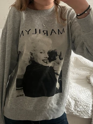 Grå sweatshirt med tryck  - Cool grå sweatshirt med stort svartvitt Marilyn Monroe-tryck framtill och texten 'MARILYN' ovanför. Tröjan har rund halsringning och långa ärmar. Perfekt för en avslappnad och trendig stil.