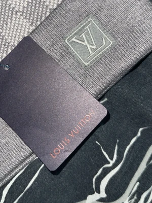 Grå Louis Vuitton mössa med rutmönster - Snygg grå mössa från Louis Vuitton med klassiskt rutmönster och LV-logga broderad framtill. Mössan har vikt kant och är tillverkad i ett mjukt, stickat material som känns lyxigt och stilrent. Perfekt accessoar för dig som gillar exklusiv streetwear.