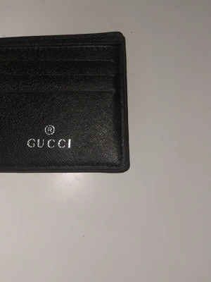 Svart korthållare från Gucci - Snygg svart korthållare från Gucci i präglat läder med plats för flera kort. Klassisk och stilren design med silverfärgad logotyp framtill. Perfekt för dig som vill ha ett lyxigt och smidigt sätt att bära dina kort.