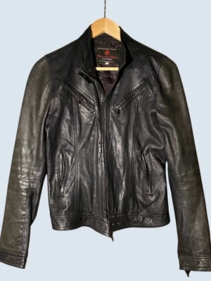 Vera pelle Svart skinnjacka med dragkedjor - Snygg svart skinnjacka från Fiorucci Jeans med flera dragkedjefickor framtill och markerad krage. Jackan har en klassisk bikerstil och detaljerade sömmar som ger en cool look. Perfekt för dig som vill ha en tidlös och edgy jacka.