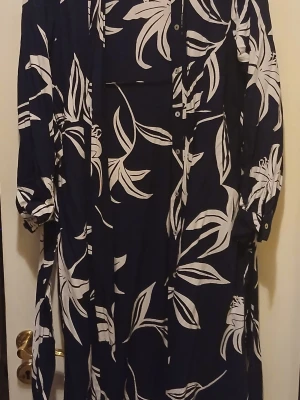 Marinblå långklänning med blommor - Snygg marinblå långklänning med stora vita blommönster och knappar framtill. Klänningen har trekvartsärm med resår vid ärmslut och ett avslappnat fall. Perfekt för dig som gillar en stilren men ändå iögonfallande look.