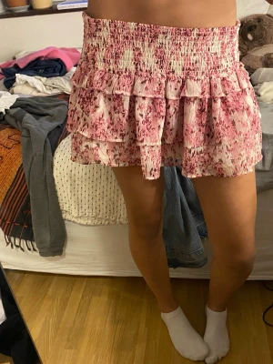 Rosa blommig volangkjol - Supersöt kort kjol med rosa blommigt mönster och volanger. Kjolen har smock i midjan för skön passform och är gjord i ett lätt, luftigt material. Perfekt för vår och sommar när du vill känna dig extra fin.