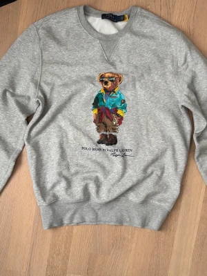 Grå Polo Bear tröja från Ralph Lauren - Säljer en grå stickad tröja från Ralph Lauren med den ikoniska Polo Bear-figuren tryckt på bröstet. Tröjan har rund halsringning och ribbade muddar vid ärmslut och nederkant. Perfekt för dig som gillar streetstyle och klassiska märken.