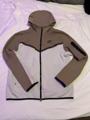 Nike Tech Fleece hoodie beige/grå - Nike Tech Fleece hoodie i beige och ljusgrått med hel dragkedja och svart Nike-logga på bröstet. Hoodien har en huva, svarta detaljer och en unik ficka med dragkedja på ena ärmen. Tillverkad i mjukt och stretchigt material för extra komfort.     MÖTAS FÖR KÖPAS (Stockholm)