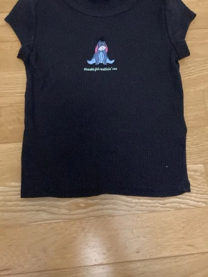 Svart ribbad topp med Nalle Puh-tryck - Kortärmad svart ribbad topp med ett tryck av Ior från Nalle Puh och texten 'thanks for noticin' me' på bröstet. Toppen har en slim passform och är perfekt för en avslappnad stil.