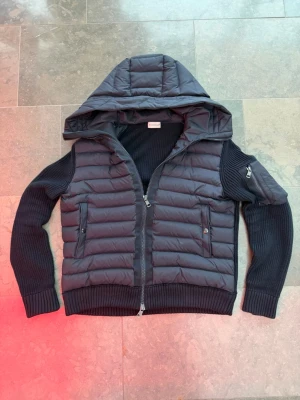 Mörkblå cardigan med huva från Moncler - Snygg svart pufferjacka från Moncler med stickade ärmar och huva. Jackan har dragkedja framtill, två fickor med knappar och en extra ficka på ärmen. Perfekt för kalla vinterdagar och riktigt trendig look.