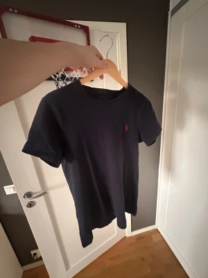 Mörkblå t-shirt från Polo Ralph Lauren - Klassisk mörkblå t-shirt från Polo Ralph Lauren med den ikoniska röda logon broderad på bröstet. T-shirten har rund hals, korta ärmar och är tillverkad i mjuk bomull. Perfekt för en clean och stilren look.