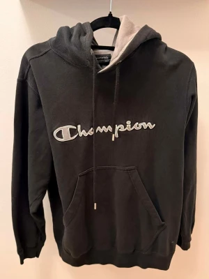 Svart Champion hoodie med logga - Snygg svart hoodie från Champion med stor broderad logga på bröstet. Hoodien har en klassisk känguruficka framtill, justerbar huva med snören och kontrastfärgad insida i huvan. Perfekt för en avslappnad streetwear-look.