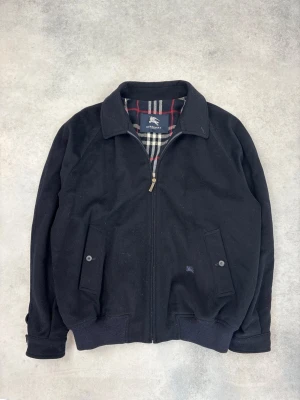 Svart bomberjacka från Burberry - Klassisk svart bomberjacka från Burberry med dragkedja framtill och två fickor med knapp. Insidan har det ikoniska rutiga Burberry-mönstret i svart, vitt och rött. Jackan har ribbade muddar och krage för en stilren look.