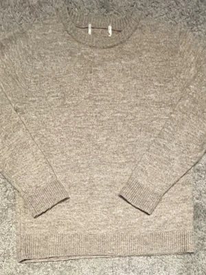 Beige stickad tröja med rund hals - Mysig beige stickad tröja med rund hals och långa ärmar. Tröjan har ribbstickade muddar vid ärmslut och nederkant, vilket ger en klassisk och stilren look. Perfekt för lager-på-lager och enkel att matcha med olika outfits.