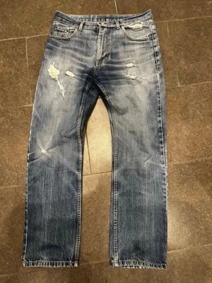 Blå slitna jeans  - Ett par blå Levi's jeans med raka ben och slitningar framtill för en cool, avslappnad look. Klassisk femficksmodell med tydliga slitdetaljer och tvättad finish. Perfekta för dig som gillar en trendig och casual stil.