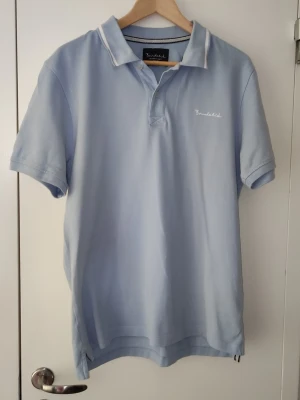 Ljusblå pikétröja från Bondelid - Snygg ljusblå pikétröja från Bondelid med klassisk krage och korta ärmar. Tröjan har vit broderad logga på bröstet och diskreta vita detaljer på kragen. Perfekt till en avslappnad och stilren look.