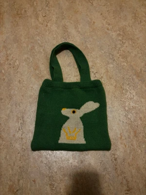 Grön stickad shoppingväska med kanin - En grön stickad shoppingväska med motiv av en vit kanin och gul krona på framsidan. Väskan har två handtag och är mjuk i materialet, perfekt för att bära med sig småsaker. Söt och unik design som sticker ut.