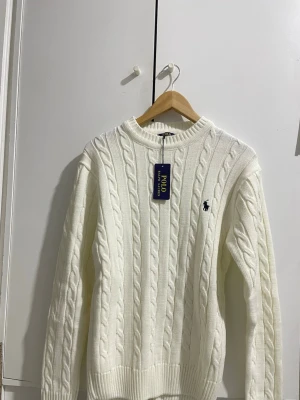 Vit kabelstickad tröja Polo Ralph Lauren - Kabelstickad vit tröja från Polo Ralph Lauren med rund halsringning och ribbade muddar. Tröjan har det klassiska broderade logotypen i marinblått på bröstet och är gjord i ett mjukt material som passar perfekt till säsongens outfits.