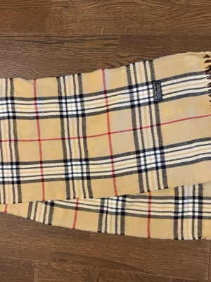 Beige Burberry Halsduk - Klassisk beige halsduk från Burberry i ull med det ikoniska rutmönstret i svart, vitt och rött. 