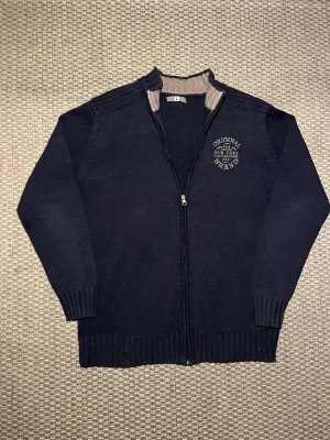 Mörkblå ziptröja  - Mörkblå stickad ziptröja  med broderad text på bröstet. Tröjan har ribbade muddar och krage med beige insida. Perfekt för lager-på-lager och har en sportig, avslappnad stil.