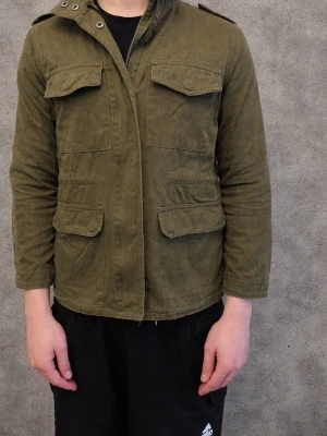 Olivgrön field jacket  - Säljer en olivgrön utilityjacka med fyra stora fickor framtill och axelklaffar. Jackan har en rak passform och är tillverkad i ett slitstarkt bomullstyg. Perfekt för dig som gillar en enkel men cool stil.