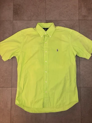Grön kortärmad skjorta från Ralph Lauren - Snygg grön skjorta från Ralph Lauren med korta ärmar och klassisk button-down krage. Skjortan har vita knappar framtill och den ikoniska lila Polo-loggan broderad på bröstet. Perfekt för dig som vill sticka ut med färg.