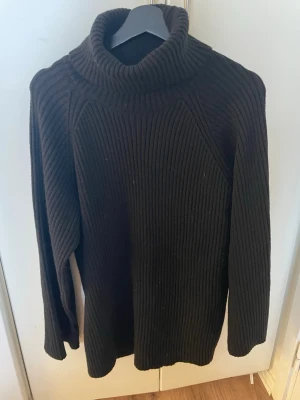 Svart polotröja i grovstickad modell - Mysig svart stickad polotröja med bred ribbstickning och hög krage. Tröjan har lång ärm och avslappnad passform, perfekt för kyliga dagar. Det är ett hål vid kragen men inget som syns när den är på se på bilden